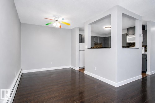 Wood-finish flooring throughout, light gray wall color, white ceiling, ceiling fan with multicolored blades, and a pass-through opening to the kitchen featuring dark cabinetry - #203 2624 Mill Woods Rd E Nw, Edmonton, AB 