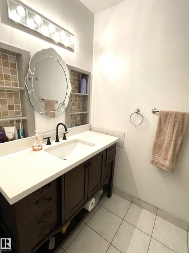 White tile flooring, dark wood-finish vanity with ample storage, white countertop with integrated sink, matte black faucet, and recessed tiled shelving - #D1 1 Garden Grove Nw, Edmonton, AB 
