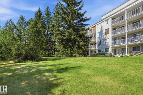 Multi-story building exterior with individual balconies, sliding glass doors, and a surrounding green lawn with mature evergreen trees - #112 4404 122 St Nw, Edmonton, AB 
