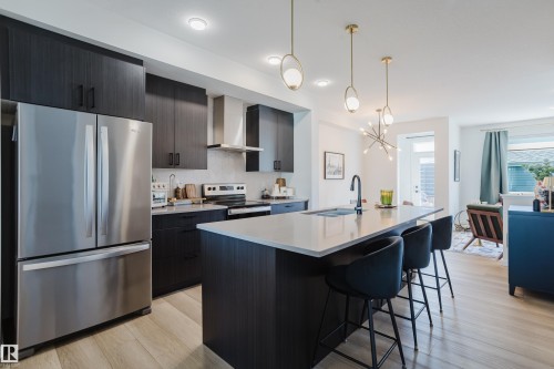 Modern kitchen featuring a large island with seating, stainless steel appliances, dark wood-finish cabinetry, light wood-finish flooring, and recessed lighting - 7017 52 Avenue, Beaumont, AB 