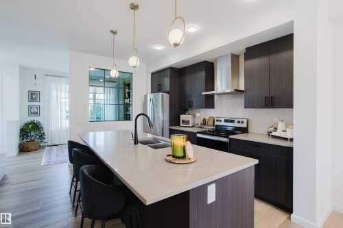Contemporary kitchen featuring dark wood-finish cabinetry, stainless steel appliances, and a light-toned countertop island with an integrated sink - 7017 52 Avenue, Beaumont, AB 