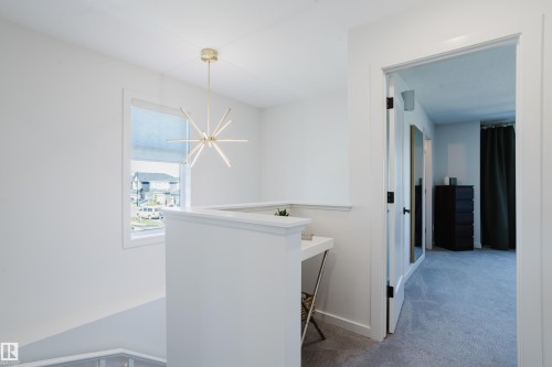 Open hallway with a modern linear chandelier, a window with roller shades, and a half-wall partition - 7017 52 Avenue, Beaumont, AB 