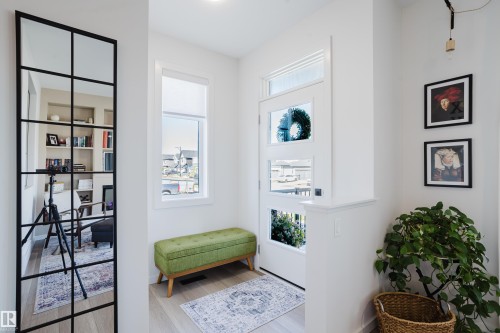 Entryway featuring light wood-finish flooring, a contemporary mirror with a black grid frame, a white panel door with multiple glass panes, and a window with a roller shade - 7017 52 Avenue, Beaumont, AB 