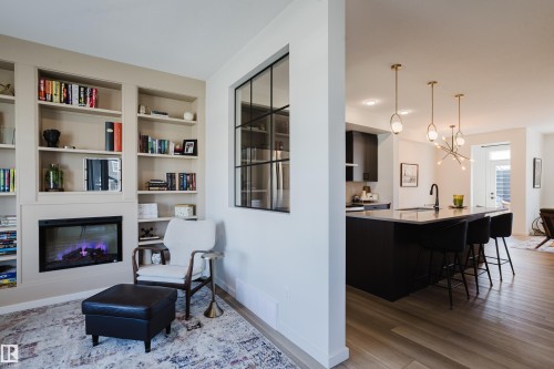 Built-in shelving with an integrated electric fireplace, wood-finish flooring, a kitchen island with seating, a black sink with a matte black faucet, and pendant lighting fixtures - 7017 52 Avenue, Beaumont, AB 