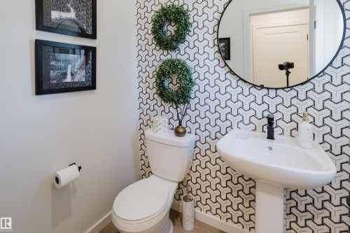 Pedestal sink with a matte black faucet, geometric pattern wall covering, round wall mirror, and modern toilet - 7017 52 Avenue, Beaumont, AB 