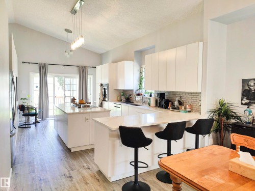 Vaulted ceiling with an architectural chandelier, wood-finish flooring, two kitchen islands with solid surface countertops, white flat-panel cabinetry, and a subway tile backsplash - 215 Peter Close Nw, Edmonton, AB 
