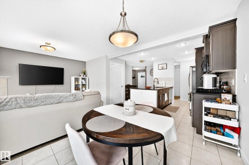 Open-concept living space featuring a dining area with a wood-finish table, a kitchen with dark cabinetry and stainless steel appliances, and tile flooring throughout - 16832 53 Street Nw, Edmonton, AB 
