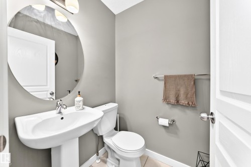 Pedestal sink with chrome faucet, circular wall mirror, wall-mounted light fixture, and tile flooring - 16832 53 Street Nw, Edmonton, AB 