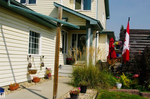 Light-colored vinyl siding exterior, concrete walkway, porch with overhead covering, partial wood fencing, and mature landscaping - 25 Hasen Close, St. Albert, AB 