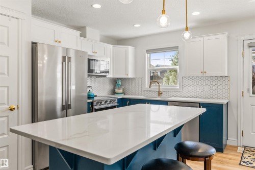 Kitchen featuring a white countertop island, stainless steel appliances, white upper cabinetry, blue lower cabinetry, and a hexagonal tile backsplash - #71 330 Bulyea Rd Nw, Edmonton, AB 