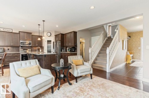 Open-concept living area featuring wood-finish flooring, a kitchen with dark cabinetry and stainless steel appliances, a kitchen island with seating, a carpeted staircase with white railings, and recessed lighting - 22 Voleta Crest, Spruce Grove, AB 