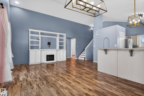 Vaulted ceilings with a decorative chandelier, wood-finish flooring, built-in entertainment center with a fireplace, and a kitchen island with a counter overhang - 13803 130A Avenue Nw, Edmonton, AB 