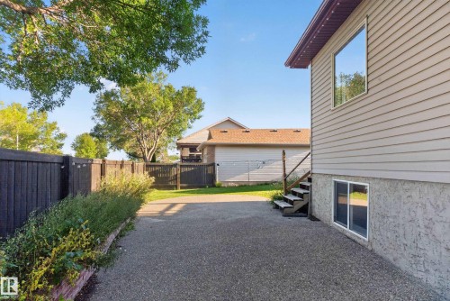 Siding exterior with multiple windows, leading to a fenced yard with mature trees and a gravel surface - 13803 130A Avenue Nw, Edmonton, AB 