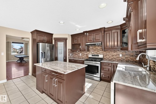 Kitchen featuring dark wood cabinetry, stainless steel appliances, light-toned tile flooring, light-toned stone countertops, and a stone-finish backsplash - 4711 152 Avenue Nw, Edmonton, AB 