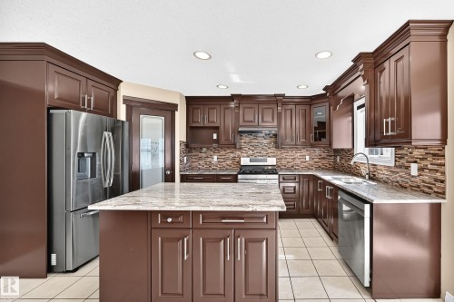 Kitchen featuring wood-finish cabinetry, stainless steel appliances, a multi-tone tile backsplash, and a center island with a light-toned countertop - 4711 152 Avenue Nw, Edmonton, AB 