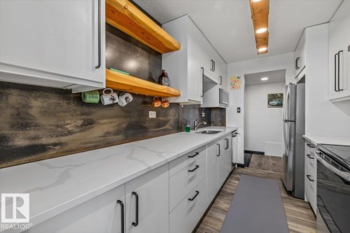 White cabinetry with matte black hardware, light-toned countertops, wood-finish flooring, stainless steel refrigerator, and integrated sink - #906 12303 Jasper Av Nw, Edmonton, AB 