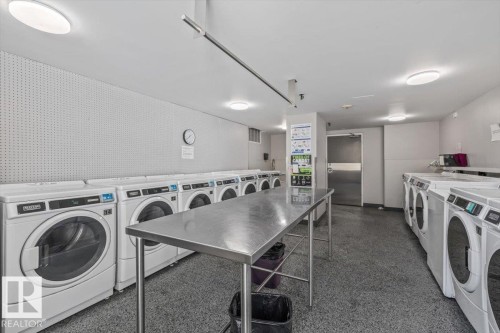Laundry facility featuring multiple front-loading washers, overhead task lighting, a central stainless steel folding table, and durable flooring - #906 12303 Jasper Av Nw, Edmonton, AB 
