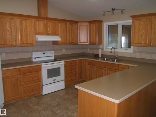 Kitchen featuring wood cabinetry, laminate countertops, tile backsplash, stainless steel sink, and tile flooring - 4825 59 Avenue, Vegreville, AB 