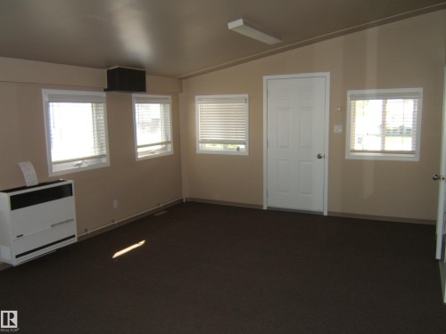 Neutral wall paint, brown carpeting, multiple windows, white door, and a wall-mounted heating unit - 4825 59 Avenue, Vegreville, AB 