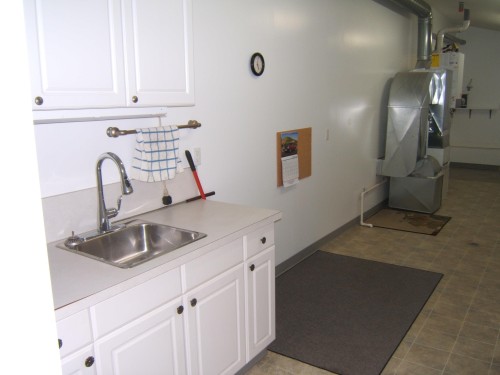 Utility space featuring white cabinetry, a stainless steel sink, a modern faucet, light-toned flooring, and exposed utility infrastructure - 4825 59 Avenue, Vegreville, AB 
