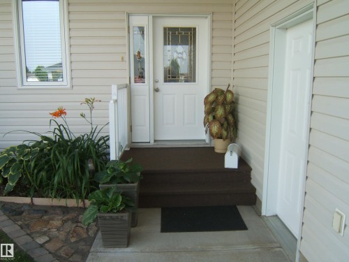 Entryway featuring a white paneled door with decorative glass inserts, an adjacent white storm door, and a brown wood-finish porch with matching steps - 4825 59 Avenue, Vegreville, AB 