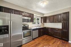 Spacious kitchen featuring dark wood cabinetry with abundant storage, stainless steel appliances including a double-wall oven and side-by-side refrigerator, white subway tile backsplash, dark countertops, and warm wood-finish flooring throughout - 