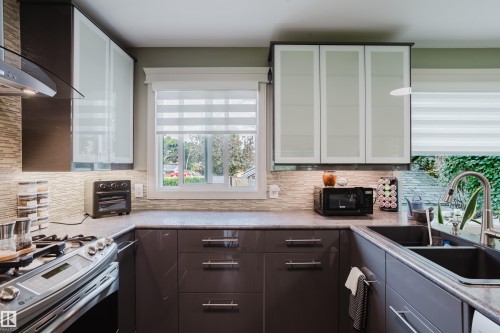 Kitchen featuring dark-finish lower cabinetry, light-finish upper cabinetry with frosted panels, a tile backsplash, and a double-basin sink with a stainless steel faucet - 14814 104 Avenue Nw, Edmonton, AB - Indoor Photo Showing Kitchen With Double Sink