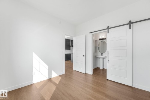 Wood-finish flooring, white walls, and a barn door leading to a bathroom featuring a pedestal sink, round mirror, and vanity light - 511 157 Avenue Ne, Edmonton, AB 