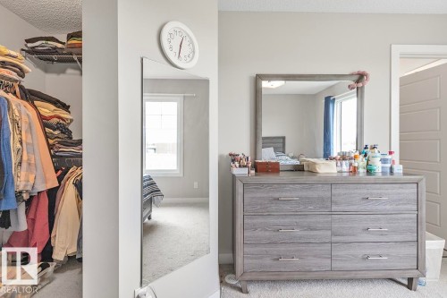 Neutral-toned room featuring a wood-finish six-drawer dresser with modern hardware, a framed wall mirror, and an adjacent walk-in closet with built-in wire shelving - #22 3025 151 Av Nw, Edmonton, AB - Indoor