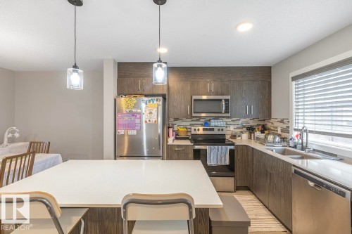 Kitchen featuring a central island with white countertop, dark wood-finish cabinetry, stainless steel appliances, and a tiled backsplash - #22 3025 151 Av Nw, Edmonton, AB - Indoor Photo Showing Kitchen With Stainless Steel Kitchen With Double Sink With Upgraded Kitchen