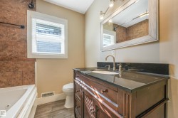 Wood-finish flooring, dark wood vanity with a black countertop, single basin sink with a polished chrome faucet, rectangular framed mirror, and a jetted bathtub with tiled surround - 