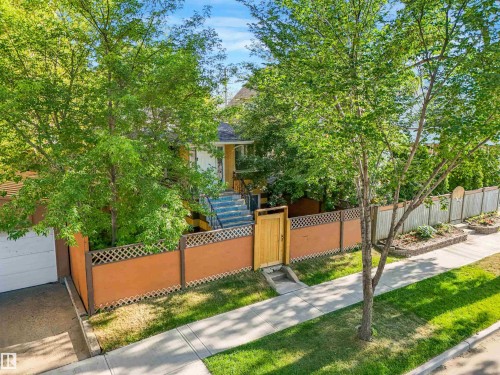 Lush mature trees framing the property, wooden gate entrance, paved sidewalk, concrete curb, and a garage door - 9347 105 Avenue Nw, Edmonton, AB 