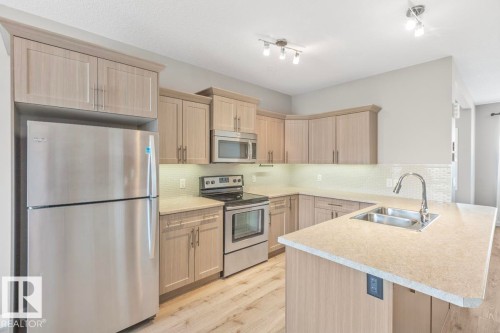 Kitchen featuring stainless steel appliances, light wood-finish cabinetry, light countertops, a tile backsplash, and wood-finish flooring - 22 Hope Common, Spruce Grove, AB 