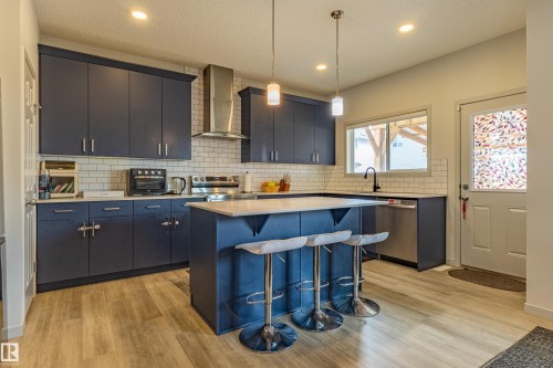 Contemporary kitchen featuring dark blue cabinetry, white subway tile backsplash, light wood-finish flooring, a central island with countertop seating, and stainless steel appliances - 41 Sequoia Bend, Fort Saskatchewan, AB 