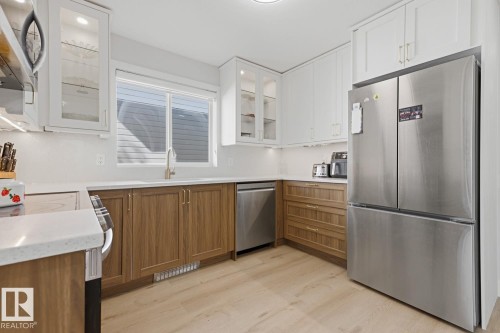 Kitchen featuring wood-finish flooring, two-tone cabinetry with upper white shaker-style doors and lower wood-finish doors, white countertops, and stainless steel appliances - 3830 22 Street Nw, Edmonton, AB 