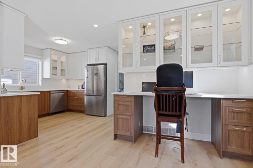 Kitchen featuring light wood-finish flooring, two-tone cabinetry with upper white shaker-style and lower wood-finish cabinets, stainless steel appliances, and a built-in desk area with glass-front upper cabinetry - 3830 22 Street Nw, Edmonton, AB 