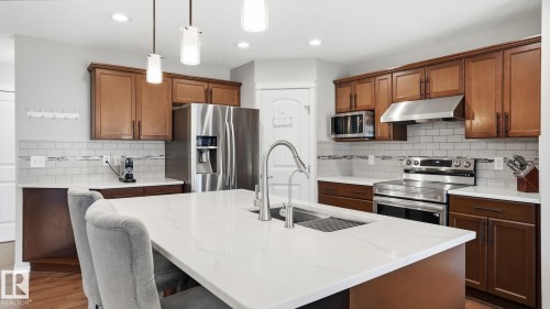 Kitchen featuring an island with a built-in sink, white countertops, wood-finish cabinetry, stainless steel appliances, and a subway tile backsplash - 4610 Westcliff Point(E) Sw, Edmonton, AB 