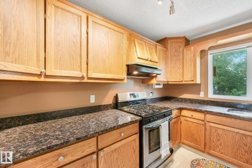 Kitchen featuring wood cabinetry, dark speckled countertops, stainless steel range with hood, and a window above the sink - 5502 45 Street, Stony Plain, AB - Indoor Photo Showing Kitchen