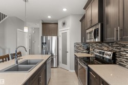 Modern kitchen featuring stainless steel appliances, dark wood-finish cabinetry, light countertops, a dual basin sink, and a tiled backsplash - 