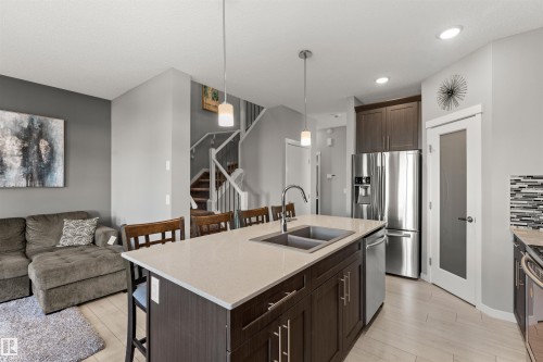 Kitchen island with light-toned countertop, stainless steel appliances, dark wood-finish cabinetry, light wood-finish flooring, and a staircase with white railings - 12218 176 Avenue Nw, Edmonton, AB 