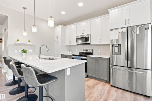 Kitchen featuring a central island with countertop seating, stainless steel appliances, two-tone cabinetry, wood-finish flooring, and pendant lighting - 8024 Orchards Green Green Sw, Edmonton, AB 
