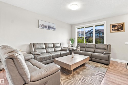 Bright living area with wood-finish flooring, a large window, light-colored walls, and a ceiling-mounted light fixture - 8024 Orchards Green Green Sw, Edmonton, AB 