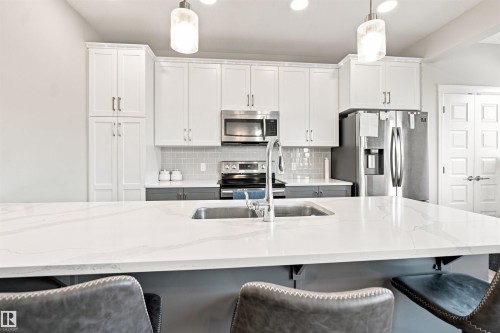 Kitchen with white upper cabinetry and gray base cabinetry, stainless steel appliances, a white island with an undermount sink, and a gray subway tile backsplash - 8024 Orchards Green Green Sw, Edmonton, AB 