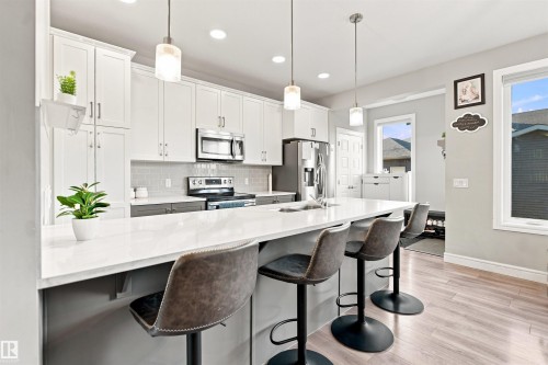 Kitchen featuring white upper cabinetry, grey lower cabinetry, stainless steel appliances, a white island with an integrated sink, and wood-finish flooring - 8024 Orchards Green Green Sw, Edmonton, AB 