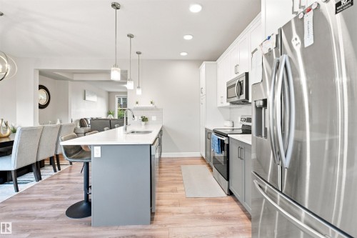 Kitchen featuring stainless steel appliances, white cabinetry, a central island with a sink, pendant lighting, and wood-finish flooring - 8024 Orchards Green Green Sw, Edmonton, AB 