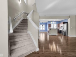 Warm wood-finish flooring extending into the kitchen, a staircase with carpeting and white railings, and a kitchen featuring stainless steel appliances - 