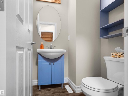 Pedestal sink with blue vanity, oval wall mirror, wall-mounted shelving, and wood-finish flooring - 5190 1B Avenue Sw, Edmonton, AB 
