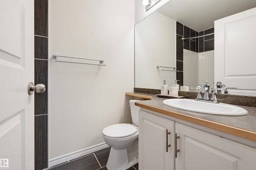 Bathroom featuring a built-in vanity with white cabinetry and an integrated sink, a large wall-mounted mirror, a two-piece toilet, dark tile flooring, and a shower with dark tile surround - #314 245 Edwards Dr Sw, Edmonton, AB - Indoor Photo Showing Bathroom