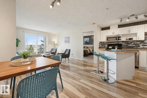 Open-concept living space featuring wood-finish flooring throughout, a modern kitchen with white cabinetry and stainless steel appliances, a dining area, and a living area with balcony access - #314 245 Edwards Dr Sw, Edmonton, AB - Indoor