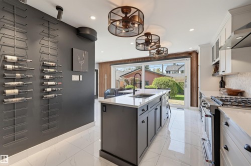 Gleaming tile flooring, a central kitchen island with integrated sink, white cabinetry with dark hardware, a stainless steel gas range, and a glass sliding door leading to the exterior - 10726 128 Street Nw, Edmonton, AB 